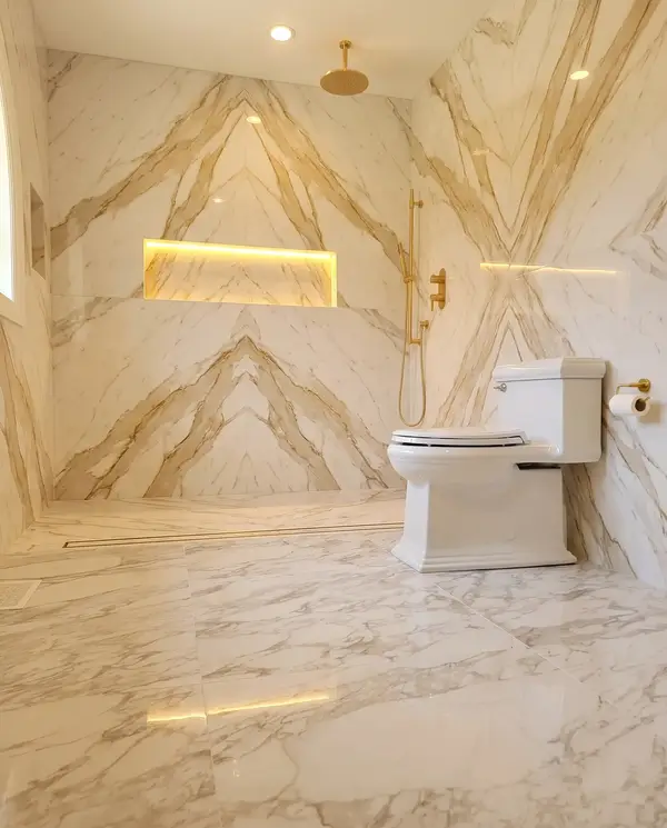 Curbless shower in bookmatched marble-look slabs with a gold rain head and lit niche