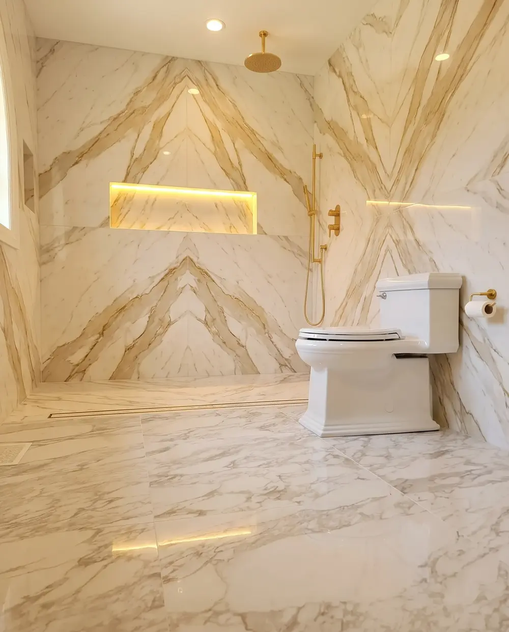 Curbless shower in bookmatched marble-look slabs with a gold rain head and lit niche