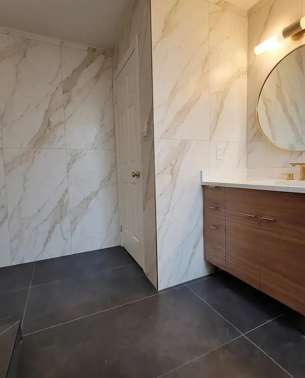 Wood vanity with a round mirror, marble-look slab walls and charcoal floor